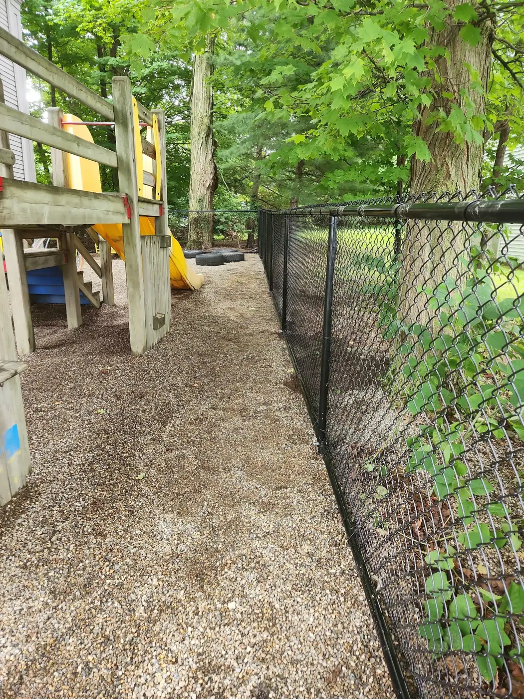 Black chain-link fence line along a playground path shaded by mature trees