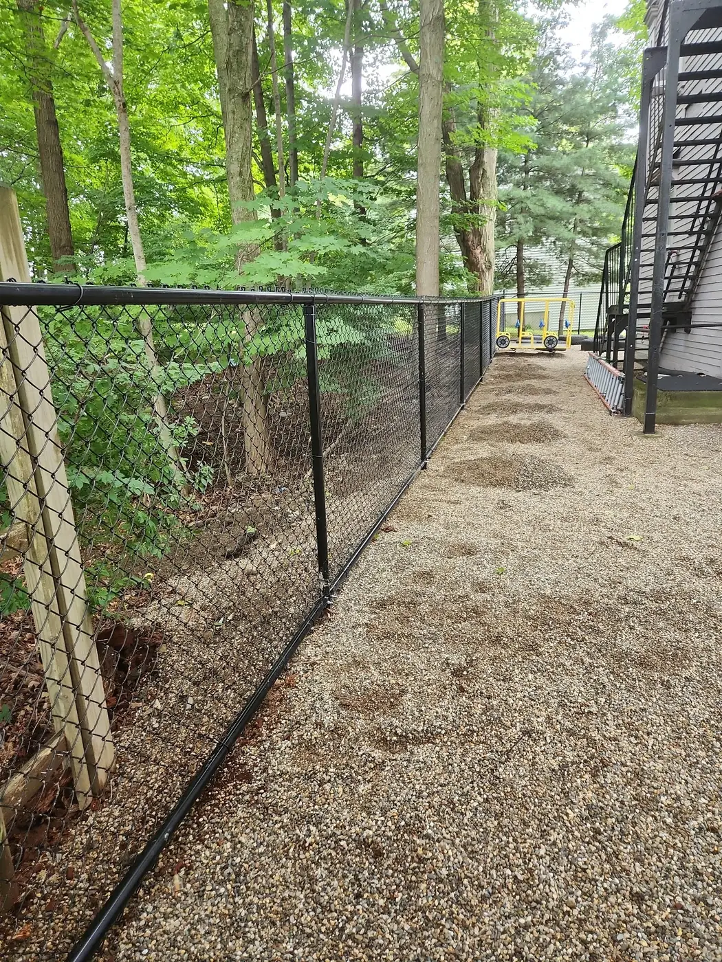 New black chain-link fence enclosing a gravel play area near the woods